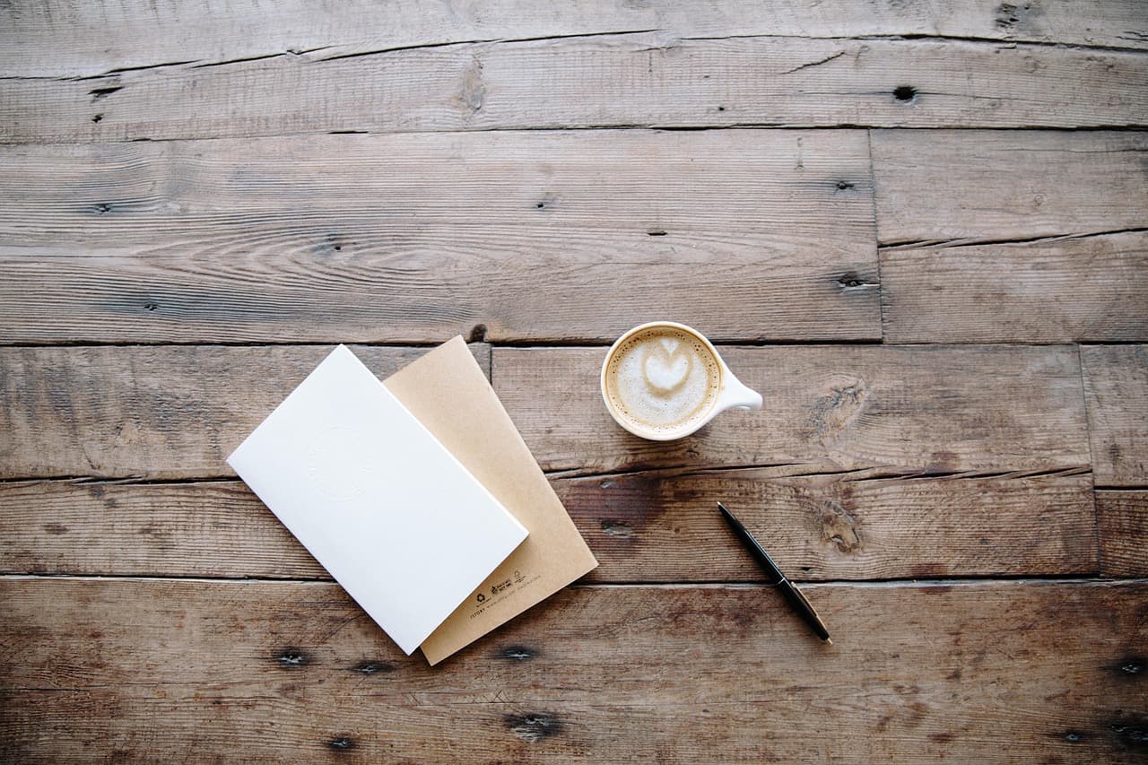 A blank letter and an envelope on a wooden table, next to a pen and a coffee
